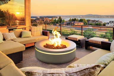 Large circular gas fire pit surrounded by cushioned patio furniture overlooking sunset views.