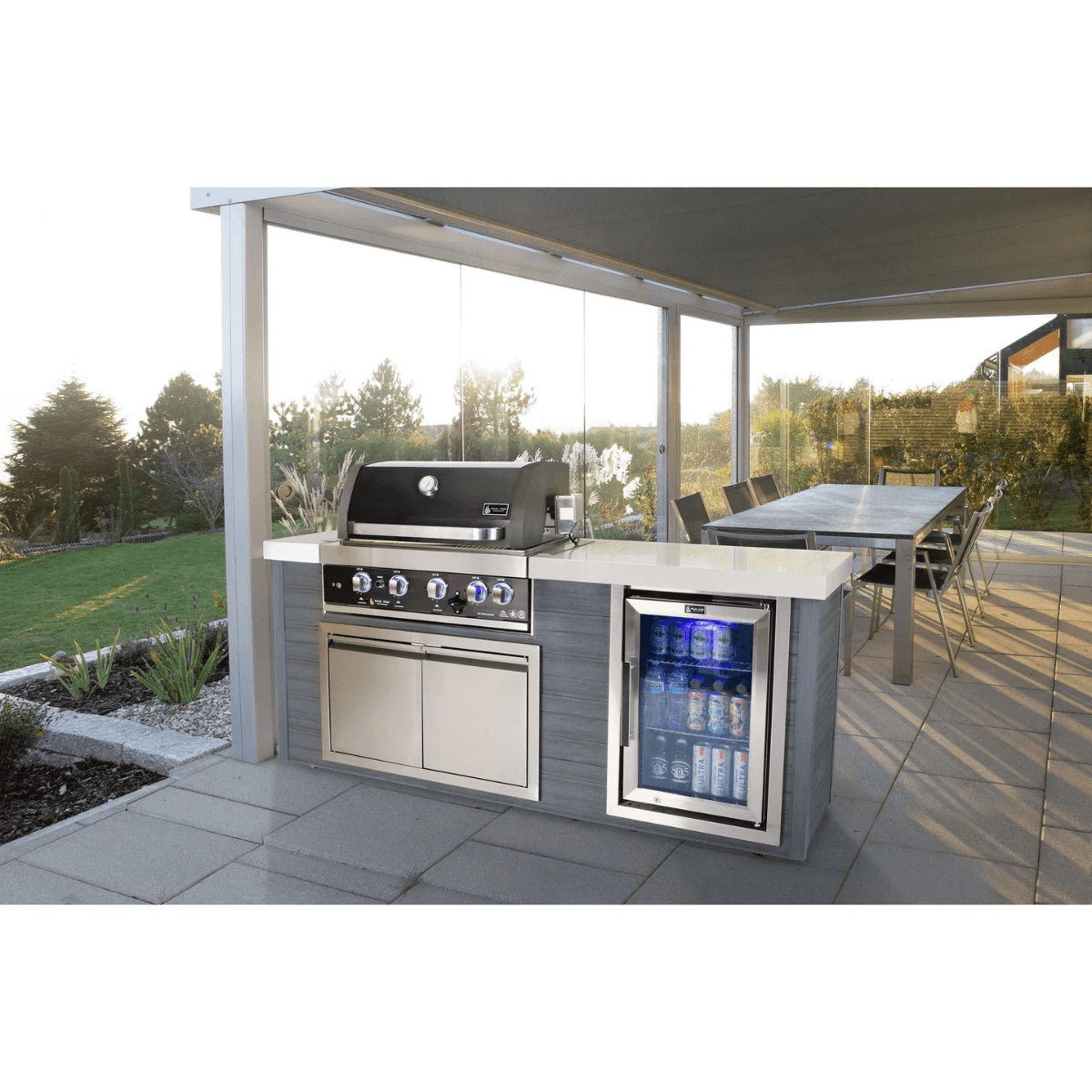 Mont Alpi 4-Burner Black Stainless Steel Art-wood Outdoor Island Grill installed in a covered outdoor kitchen, featuring a black stainless steel hood, beverage fridge, and spacious dining area.