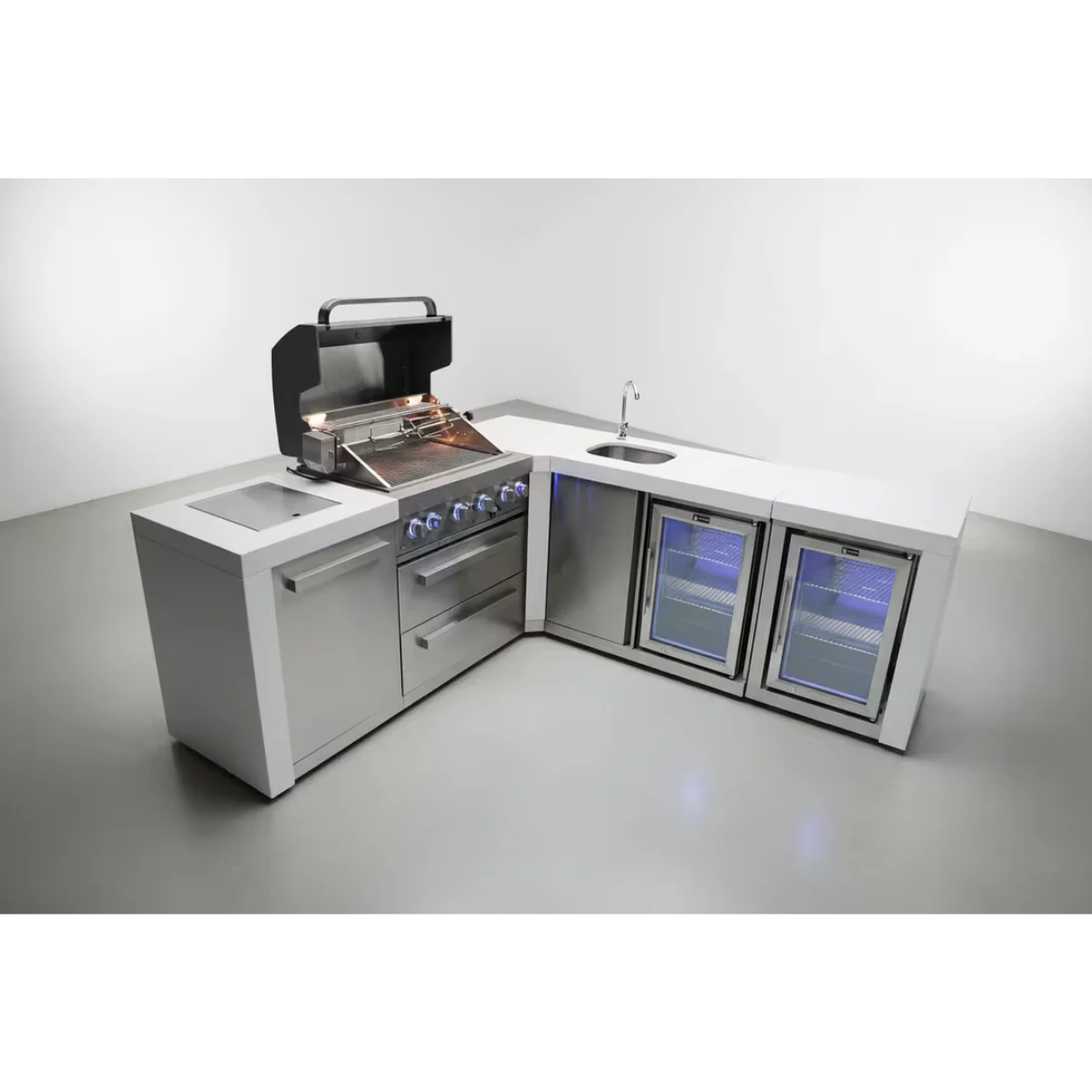 Angled view of the Mont Alpi 400 Deluxe Island with 90-degree corner, built-in gas grill, sink, beverage center, and fridge cabinet with illuminated glass doors.