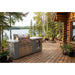 Mont Alpi 6-Burner Black Stainless Steel Art-wood Outdoor Island Grill on a lakeside cabin deck, featuring a built-in beverage fridge, storage cabinet, and scenic woodland backdrop.