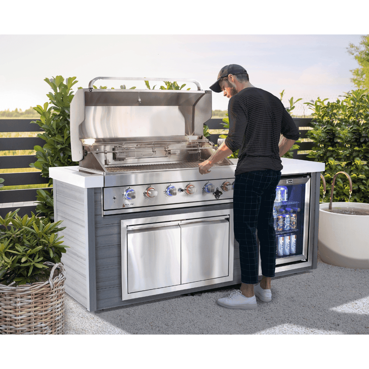 Person grilling at the Mont Alpi 6-Burner Stainless Steel Art-wood Outdoor Island Grill with open hood, integrated beverage fridge, storage cabinet, and modern outdoor garden setting.