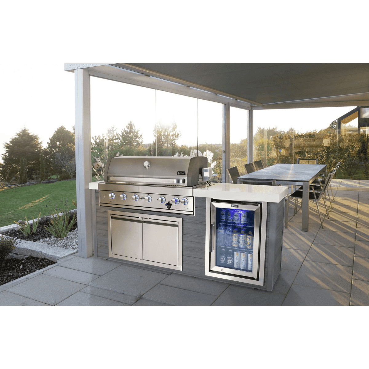Mont Alpi 6-Burner Stainless Steel Art-wood Outdoor Island Grill installed under a covered patio, featuring a built-in fridge, storage cabinet, and spacious outdoor dining area.