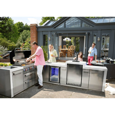 Outdoor entertaining scene featuring the Mont Alpi 805 Deluxe Island with 90-degree corner, kegerator, beverage center, and guests gathered around the grill.