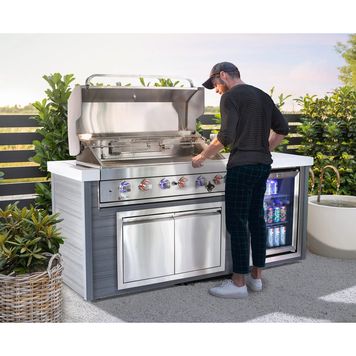 Outdoor entertaining area featuring a Mont Alpi deluxe grill island with an open stainless steel barbecue, built-in beverage fridge, storage cabinet, and LED-lit control knobs while a person prepares food on a sunny patio.