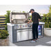 Outdoor entertaining area featuring a Mont Alpi deluxe grill island with an open stainless steel barbecue, built-in beverage fridge, storage cabinet, and LED-lit control knobs while a person prepares food on a sunny patio.