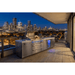 Stainless steel Mont Alpi 805 Deluxe Island with kegerator on a rooftop terrace overlooking a city skyline at night.