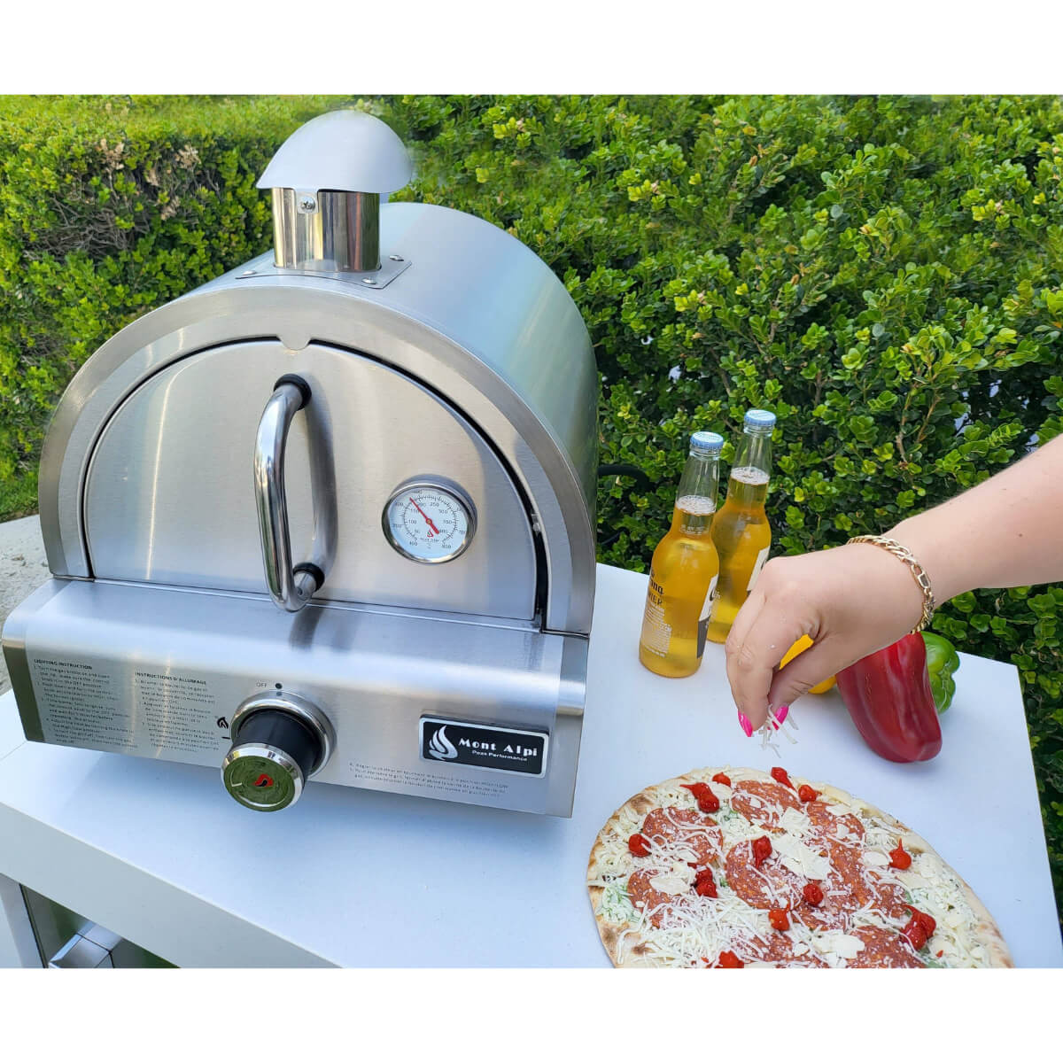 Stainless steel Mont Alpi portable pizza oven in an outdoor setting with a freshly prepared pizza and ingredients nearby.