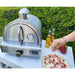 Stainless steel Mont Alpi portable pizza oven in an outdoor setting with a freshly prepared pizza and ingredients nearby.