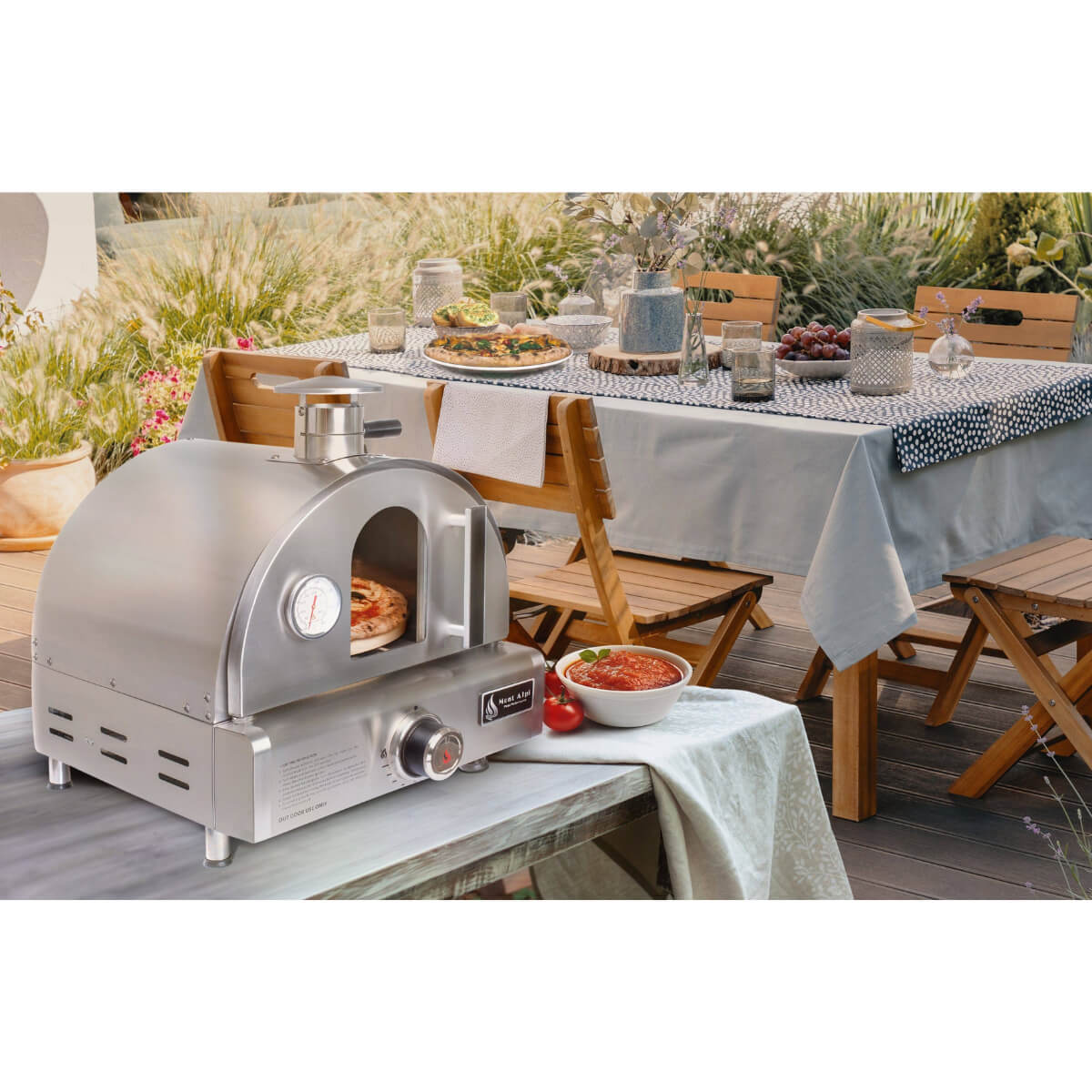 Mont Alpi portable pizza oven displayed on an outdoor dining table beside pizza ingredients, ready for alfresco cooking and entertaining.