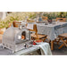 Mont Alpi portable pizza oven displayed on an outdoor dining table beside pizza ingredients, ready for alfresco cooking and entertaining.