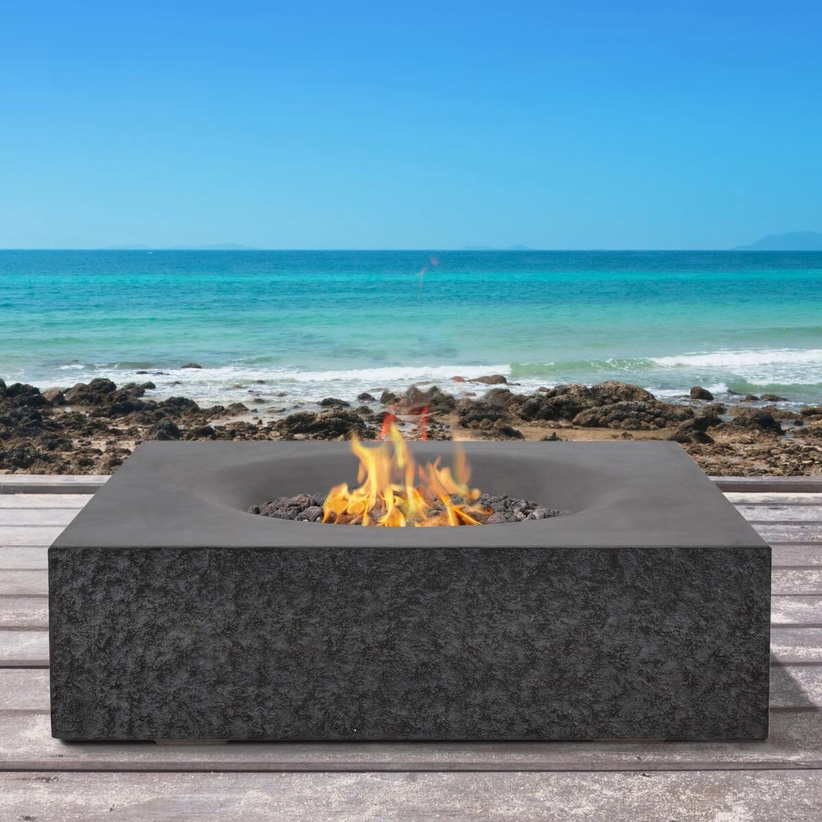 Dark grey Pyromania Monument fire table with active flames overlooking the ocean.